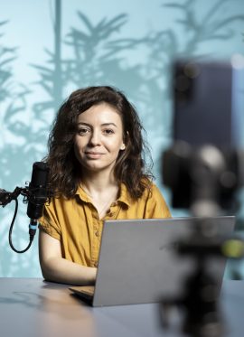Smiling vlogger sitting in front of camera, reading from laptop screen, using professional microphone. Cheerful influencer scrolling through text on notebook, addressing their audience