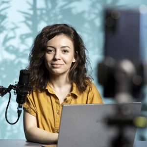 Smiling vlogger sitting in front of camera, reading from laptop screen, using professional microphone. Cheerful influencer scrolling through text on notebook, addressing their audience